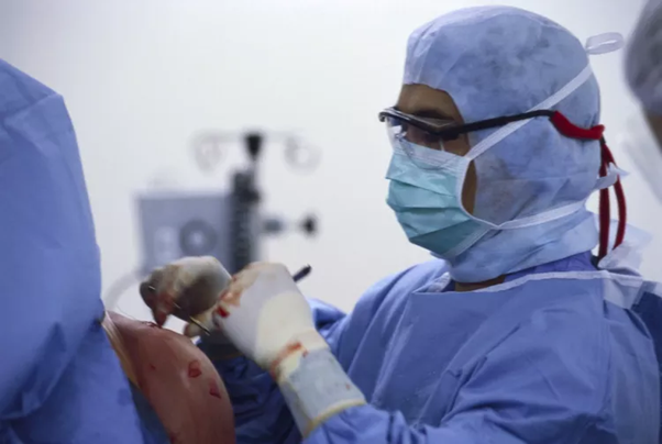 Surgeon in blue scrubs, mask and goggles performing surgery on a patient in an operating room.