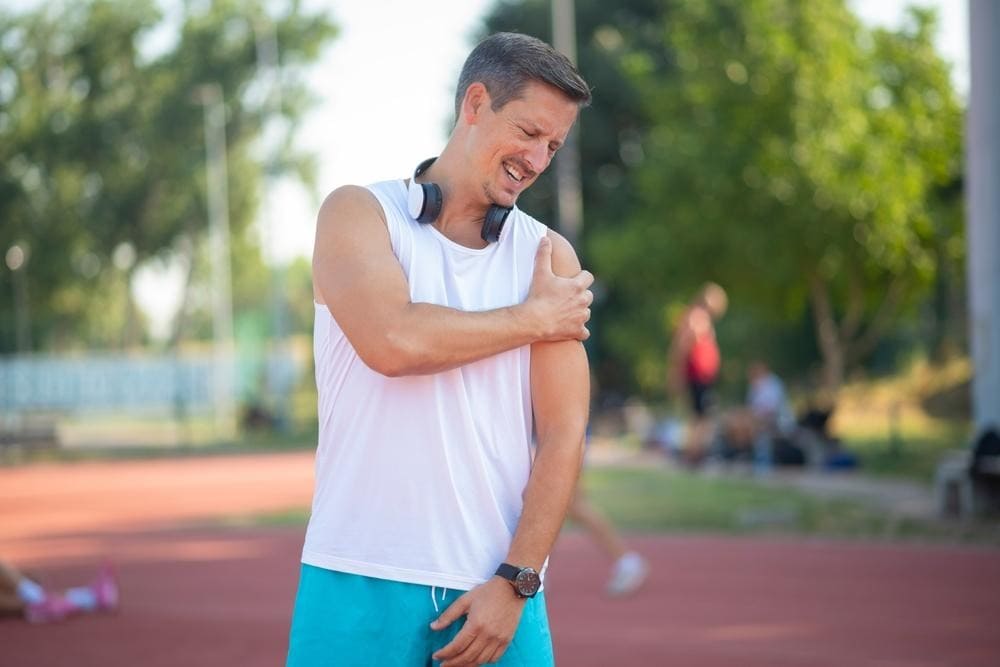 Happy man in a white sleeveless shirt and blue shorts stretches his shoulder on an outdoor running track, with headphones around his neck.