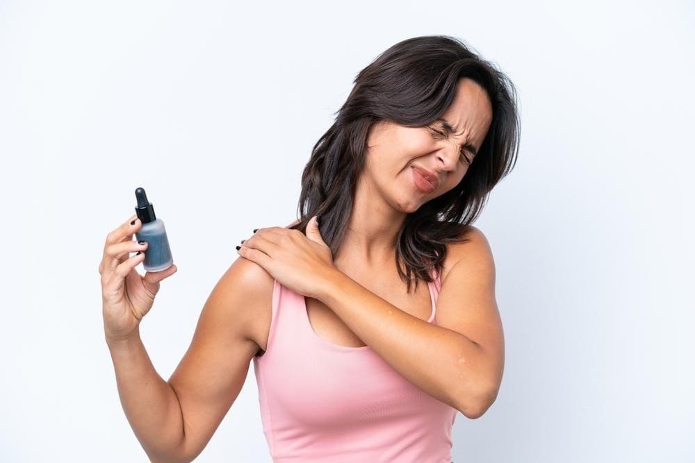 Woman in a pink tank top grimaces in pain while holding a dropper bottle and touching her shoulder