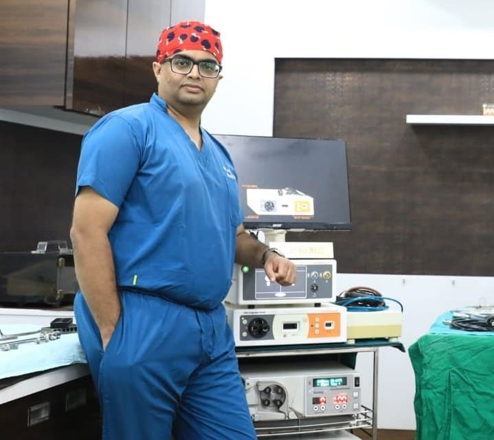 p7 Medical professional in blue scrubs and a red surgical cap standing in a hospital operating room beside medical equipment and monitors.